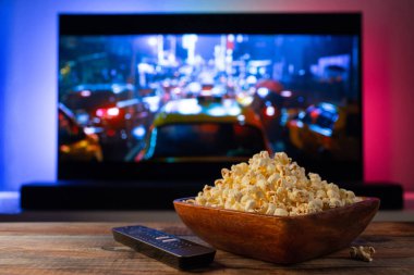 Arkaplanda bir kase patlamış mısır ve uzaktan kumanda var. Televizyon çalışıyor. Akşam sıcacık, evde film ya da TV dizisi seyrediyor.