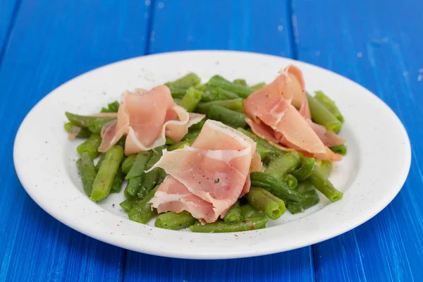 Green beans with prosciutto on white plate on wooden background — Stock Photo, Image