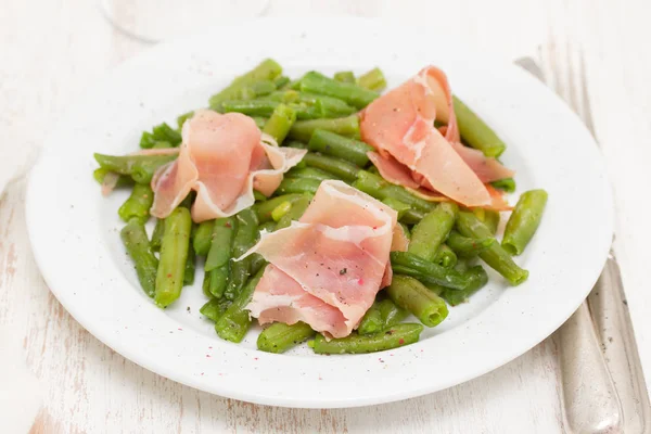 Green beans with prosciutto on white plate on wooden background — Stock Photo, Image