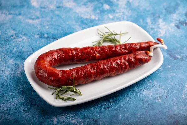 Salsiccia affumicata con rosmarino su piatto bianco su fondo blu — Foto Stock