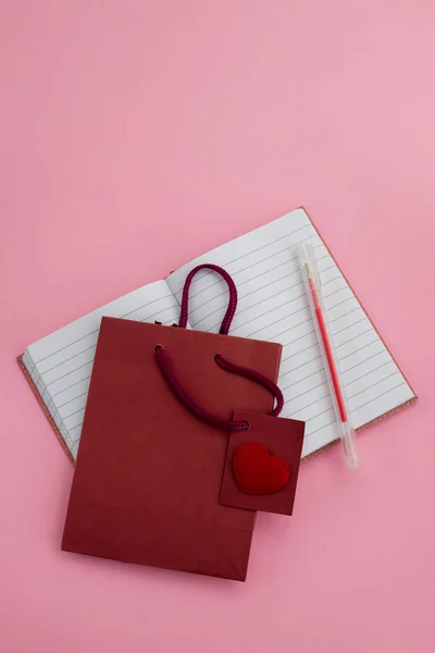 Corazón con una pequeña bolsa de papel y un cuaderno con un bolígrafo en respaldo rosa —  Fotos de Stock