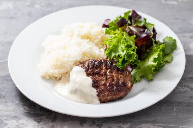 Soslu ızgara hamburger, haşlanmış pirinç ve beyaz tabakta taze salata.
