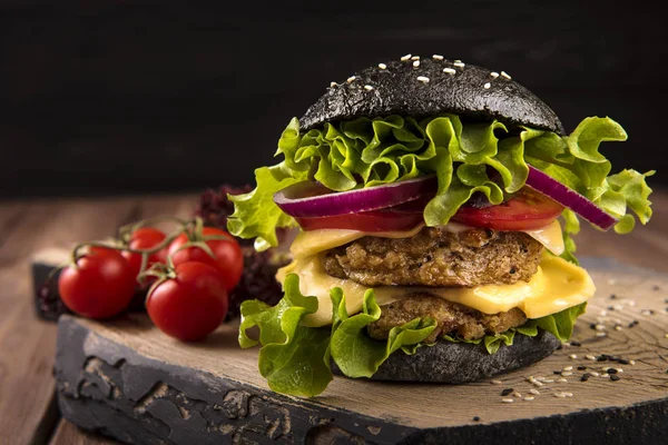 Veganer schwarzer Burger mit zwei Kichererbsenschnitzeln, Tomaten, Käse, Zwiebeln und Salat auf Holztisch, dunkler Hintergrund — Stockfoto