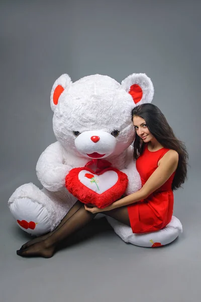 Ung vacker flicka i röd klänning med stor nallebjörn mjuk leksak glada leende och spelar på grå bakgrund — Stockfoto