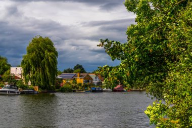 Geniş nehir ve evleri sahilinde demirli deniz araçları, yeşil vegetati