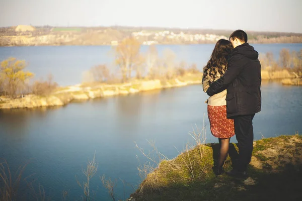 Man en vrouw aan de oever van het meer met rotsachtige kusten, in het vroege voorjaar. Silhouetten van liefhebbers die gaan in het water op de achtergrond van de zonsondergang op een meer. — Stockfoto