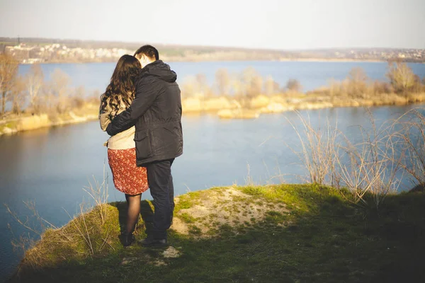 Man en vrouw aan de oever van het meer met rotsachtige kusten, in het vroege voorjaar. Silhouetten van liefhebbers die gaan in het water op de achtergrond van de zonsondergang op een meer. — Stockfoto