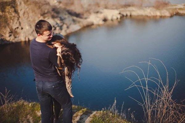 Esposo y esposa en la orilla del lago con costas rocosas, principios de primavera. Siluetas de amantes que entran en el agua sobre el fondo de la puesta de sol sobre un lago . — Foto de Stock