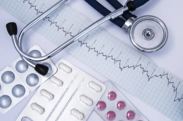 Composición o idea para la dirección médica de la medicina interna: un estetoscopio, un electrocardiograma (ECG o ECG) y un conjunto de píldoras blancas y púrpuras en blisters acostados en la mesa médica blanca —  Fotos de Stock