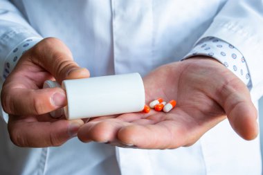Doctor or medical professional holds plastic container with orange capsules and pours them into other hand. Concept of prescribing medical drug therapy in form of pharmaceuticals such as antibiotics clipart