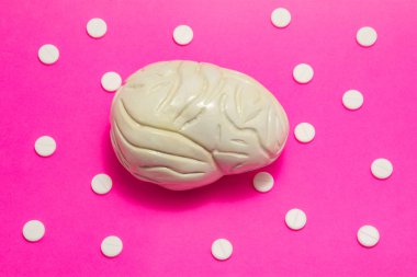 Beynin 3 boyutlu anatomik modeli pembe arka planda, süs puanı olarak beyaz haplarla çevrili. Tıbbi konsept farmakolojik masa tedavisi beyin ve nöroloji hastalıkları ilaç terapisi