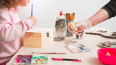 Küçük kız ahşap bir kutuya akrilik boyalı beyaz bir tek boynuzlu at çiziyor..