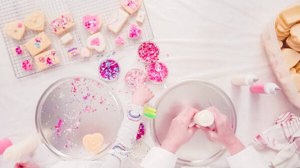 Flat lay. Step by step. Little girl decarting sugar cookies with royal icing and sprinkles for Valentine's Day.