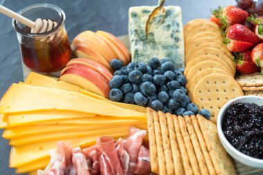 Aperatif olarak sunulan tahtanın üzerinde gurme peyniri, kraker ve meyve var..