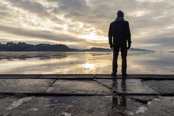 Man standing at the shore, looking at the calm sea. Reflections of the man in the ice on the ground. Mist and fog. Hamresanden, Kristiansand, Norway