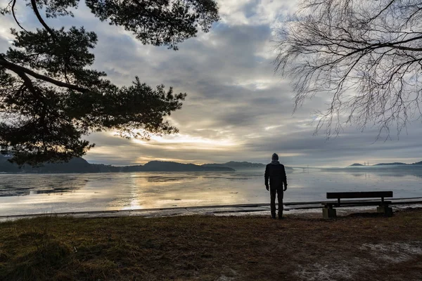 Homme debout seul à côté d'un banc au bord de la mer, regardant l'eau calme et la brume et le brouillard. Hamresanden, Kristiasand, Norvège — Photo