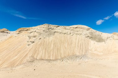 Mavi gökyüzü ve beyaz bulutlar gibi bir dağ tepe görünüyor arka planda büyük bir kum çukurunda kum