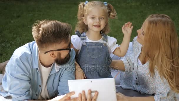 Ritratto di famiglia di felice ragazza e mamma e papà divertirsi sull'erba in soleggiata giornata estiva e tablet gioco — Video Stock
