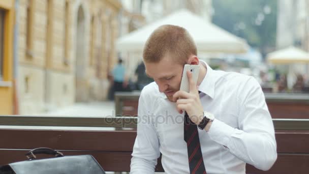 Молодий бізнесмен, сидячи на лавці, працює за домовленістю і розмовляє по мобільному телефону — стокове відео