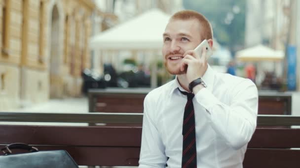 Joven hombre de negocios sentado en la banca trabajando en el acuerdo y hablando por teléfono móvil — Vídeos de Stock