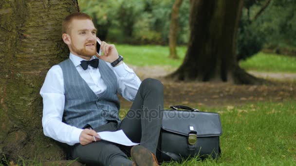 Joven hombre de negocios está sentado bajo el árbol — Vídeo de stock