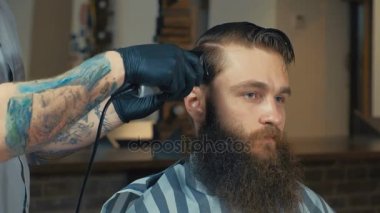 Eşsiz sakallı ve dövmeli berber berber dükkanında müşterisinin saçını kesiyor. Tarak ve saç makası kullanıyor..