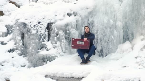 Щасливий молодий чоловік грає на гармонії на замерзлому водоспаді — стокове відео