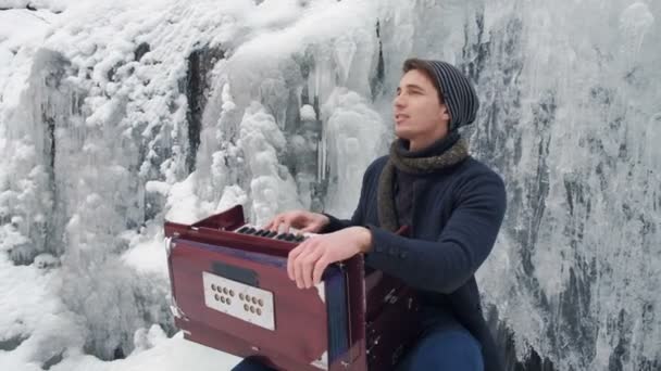 Щасливий молодий чоловік грає на гармонії на замерзлому водоспаді — стокове відео