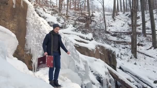 Щасливий молодий чоловік грає на гармонії на замерзлому водоспаді — стокове відео