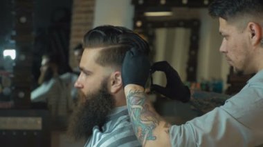 Eşsiz sakallı ve dövmeli berber berber dükkanında müşterisinin saçını kesiyor. Tarak ve saç makası kullanıyor..