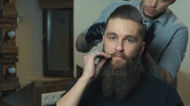 Çekici koyu saçlı adam beyaz gömlek bir saç kesimi saç kurutma makinesi ve adam doğru el bıyık ve kameraya seyir için saç fırçası ile yapıyor.