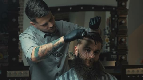 Um barbeiro sem paralelo com barba e uma tatuagem está a cortar o cabelo do seu cliente na barbearia. Ele está usando um pente de corte e um cortador de cabelo. — Vídeo de Stock