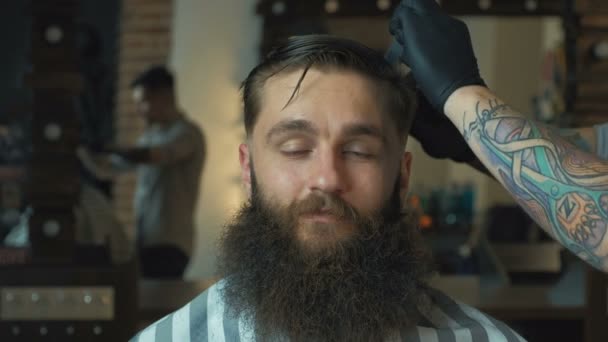 Fryzjer, ciemne włosy i tatuaż na sobie białą koszulę i czarny rękawiczki robi fryzura dla klienta z nożyczkami w fryzjera, światła w tle, portret. — Wideo stockowe