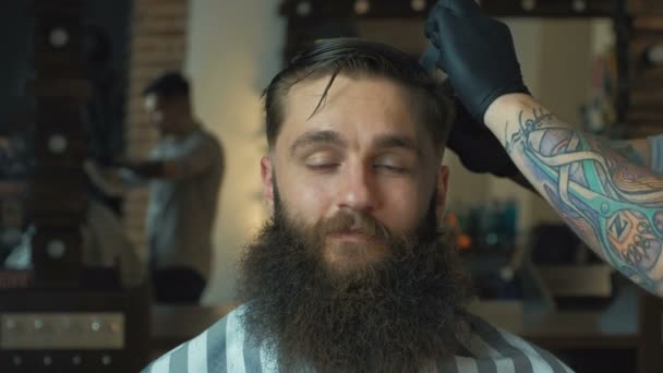 Barbeiro com cabelo escuro e tatuagem vestindo camisa branca e luvas pretas fazendo um corte de cabelo para o cliente com tesoura na barbearia, luzes no fundo, retrato . — Vídeo de Stock