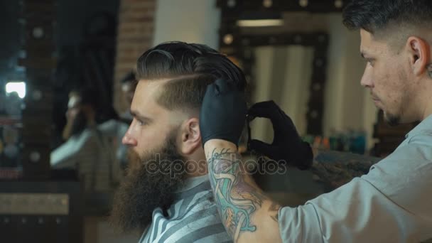 Un coiffeur sans pareil avec une barbe et un tatouage coupe les cheveux de son client dans le salon de coiffure. Il utilise un peigne de coupe et une tondeuse. — Video