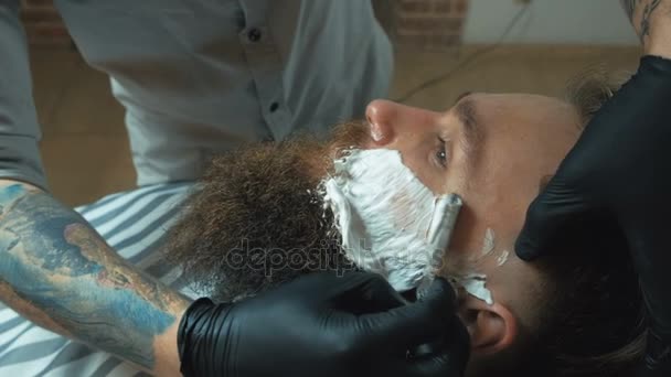 Homem bonito com tatuagem vestindo camisa branca e luvas fazendo forma de barba com navalha para homem com cabelo preto na barbearia, retrato . — Vídeo de Stock