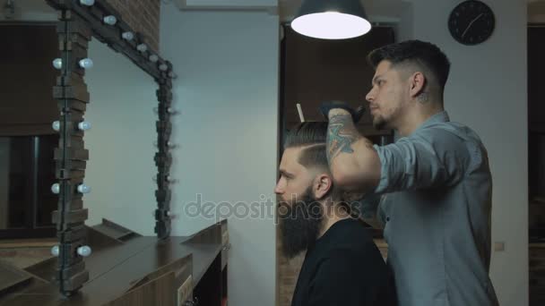 Peluquero desigual con una barba y un tatuaje está secando el cabello de su cliente en una capa de corte de pelo azul en la barbería. Está usando un secador de pelo negro y un cepillo. Horizontal . — Vídeos de Stock