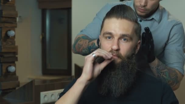 Atractivo hombre de pelo oscuro con camisa blanca haciendo un corte de pelo con secador de pelo y cepillo de pelo para el hombre bigote mano correcta y mirando a la cámara . — Vídeos de Stock