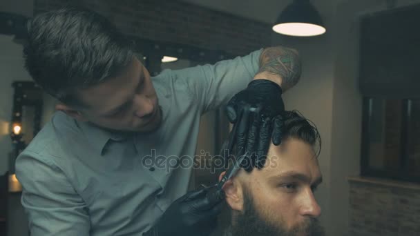 Fryzjer z ciemnymi włosami i tatuaż na sobie białą koszulę i czarny rękawiczki robi fryzura dla klienta brutalny nożyczkami barber Shop — Wideo stockowe