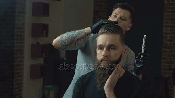 Nuevo peinado. Vista lateral del joven barbudo que se arregla en la peluquería con secador de pelo mientras está sentado en la silla en la barbería — Vídeos de Stock