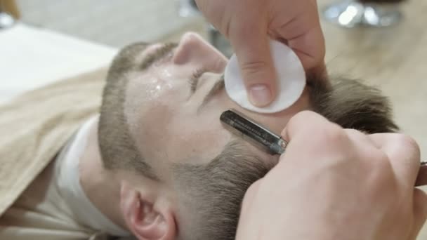 Close-up portrait of handsome young man getting beard shaving with straight razor. Focus on the blade — Stock Video