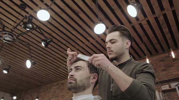 Mestre corta cabelo e barba de homens na barbearia, cabeleireiro faz penteado para um jovem — Vídeo de Stock