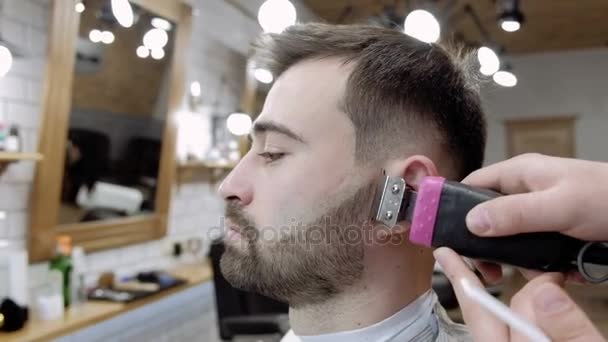 Mestre corta cabelo e barba de homens na barbearia, cabeleireiro faz penteado para um jovem — Vídeo de Stock