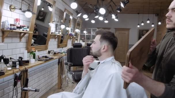 El barbero sostiene un espejo y muestra al cliente real de vuelta en la barbería — Vídeo de stock