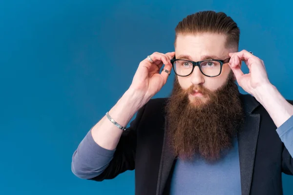 Att ta livet på allvar. Porträtt av stilig ung man i formalwear justera glasögonen stående mot blå bakgrund. — Stockfoto