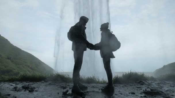 Пара, що стоїть на пагорбі біля водоспаду. Силует пари тримає руки — стокове відео