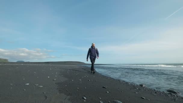 Молодой человек идет по черному песчаному пляжу на горе. Во время отпуска мужчина в теплой одежде. Человек на берегу прогулки — стоковое видео