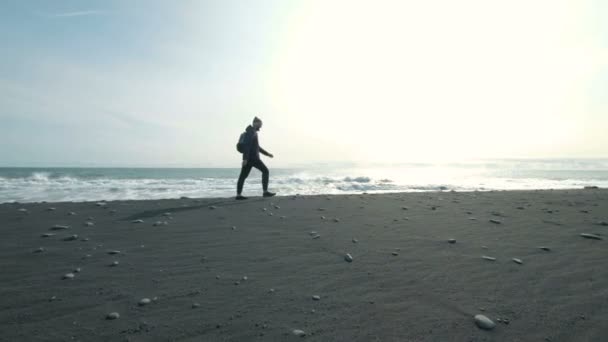 Junger Mann, der an einem schwarzen Sandstrand auf einem Berg spaziert. Im Urlaub ist der Mann warm angezogen. eine Person befindet sich am Ufer eines Spaziergangs — Stockvideo