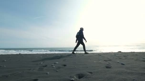 Fiatal férfi gyaloglás-ra egy fekete homokos strandjától, a hegyen. Nyaralás alatt az ember van a meleg ruhát. Egy személy van egy séta a parton — Stock videók