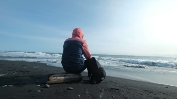 Молодой человек сидит на черном пляже и смотрит на море — стоковое видео
